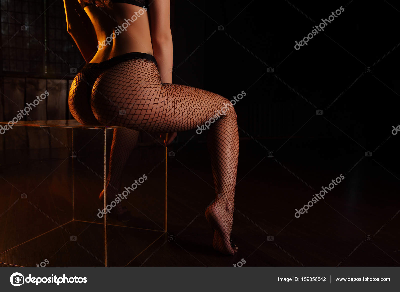 Hips, legs and ass. woman in black lingerie and fishnet tights sitting on  transparent cube in dark studio. Lighted with orange from side Stock Photo  by ©fenixlive 159356842