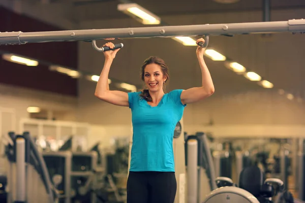 Strong woman in blue t-shirt and black pants exercising in a gym - doing pull-ups.