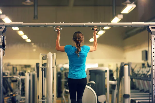 Strong woman in blue t-shirt and black pants exercising in a gym - doing pull-ups..