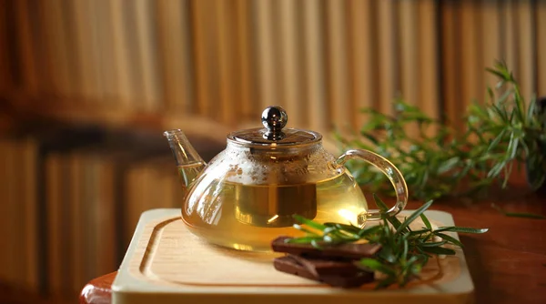 Teekanne mit Kräutertee auf Holzschneidebrett mit Rosmarin und Schokolade in der Nähe, Bücher im Hintergrund, gemütliches Interieur zu Hause — Stockfoto