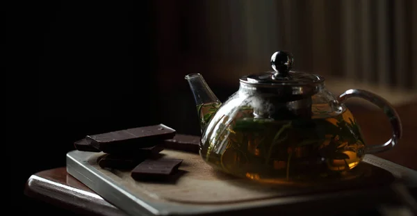 Teekanne mit Kräutertee auf Holzschneidebrett mit Schokolade in der Nähe, Bücher im Hintergrund, gemütlicher Abend zu Hause — Stockfoto