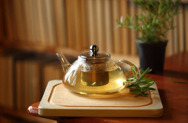 Чайник с травой чай на деревянной доске резки с розмарином рядом, книги на заднем плане, домашний уютный интерьер — стоковое фото