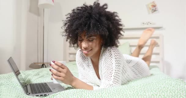 Αισθησιακή νεαρή αφρικανική αμερικανική γυναίκα με Άφρο hairstyle χαλαρωτικό στο υπνοδωμάτιο, χρησιμοποιώντας φορητό υπολογιστή και πόσιμο ζεστό καφέ. — Αρχείο Βίντεο