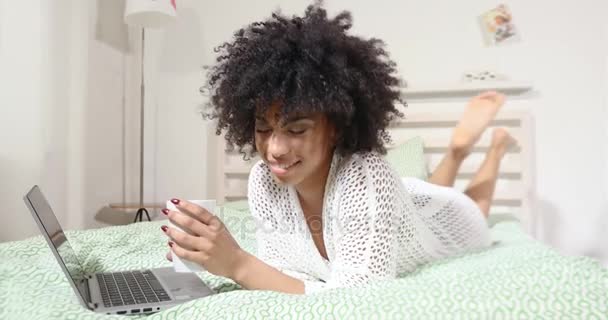 Sensuele jonge african american vrouw met afro kapsel ontspannen in slaapkamer, met behulp van de laptop en het drinken van hete koffie. — Stockvideo