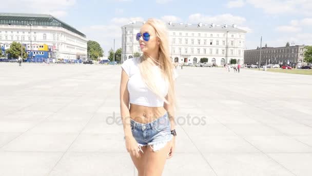 Bela jovem caucasiana caminhando na cidade, vestindo óculos de sol elegantes e jeans curtos, dia ensolarado de verão . — Vídeo de Stock