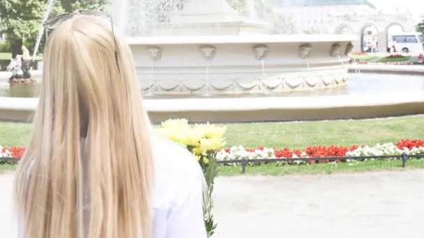 Hermosa mujer rubia caucásica sonriendo a la cámara, posando con flores en el parque de la ciudad . — Vídeo de stock