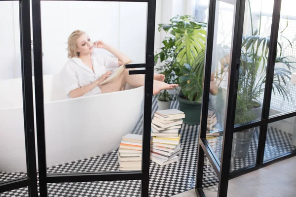 Schönes blondes Mädchen liest ein Buch im Badezimmer. — Stockfoto