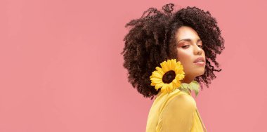 Pembe pastel stüdyo arka planında sarı ayçiçeğiyle poz veren güzel afro kadın. Kadınlar ve anneler günü kavramı. Bahar, yaz hisleri. Kız gücü.