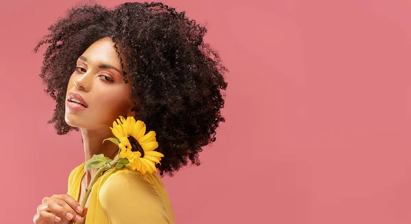 Beautiful afro woman posing with yellow sunflower on pink pastel studio background. Concept of women's and mother's day. Spring, summer feelings. Girl power.