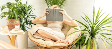 Rahatlayan, yeşil ev bitkileriyle dolu rahat bir odada kitap okuyan bir kadın. Boho ev dekorasyonu. Kitap kurdu kavramı.