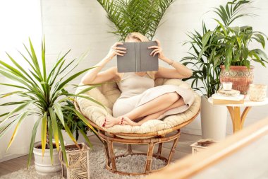 Rahatlayan, yeşil ev bitkileriyle dolu rahat bir odada kitap okuyan bir kadın. Boho ev dekorasyonu. Kitap kurdu kavramı.