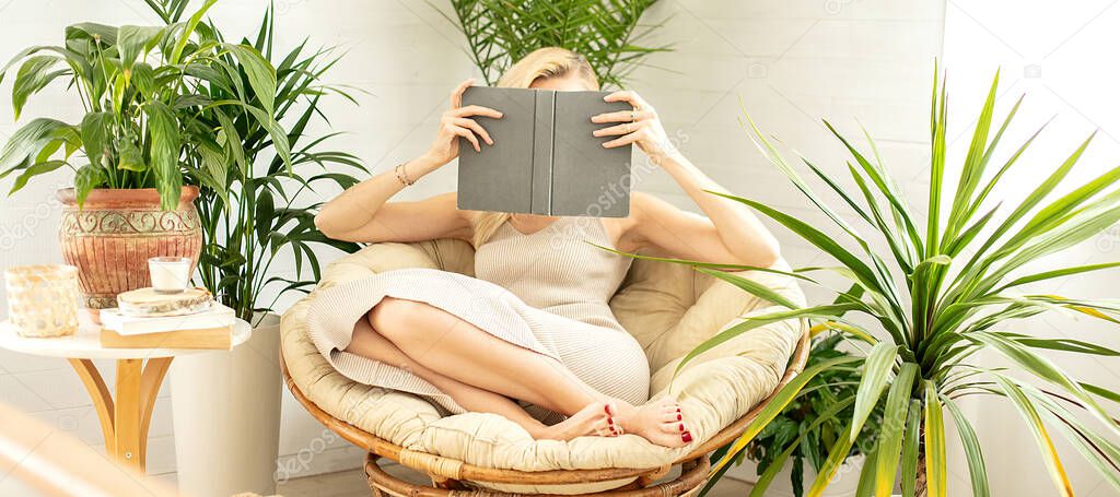 Woman relaxing, reading a book in cozy room with green home plants. Boho home decor. Bookworm concept.