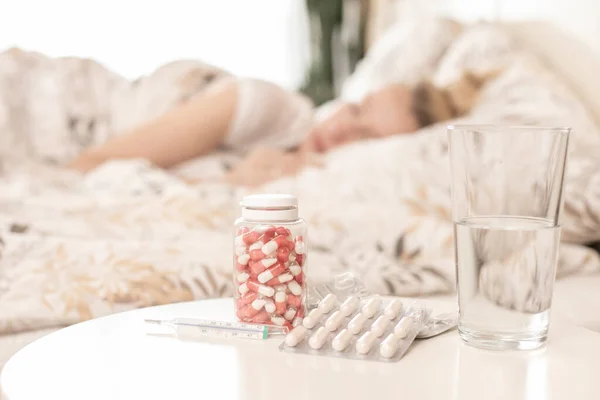 Sick woman lying in bed, sleeping. Glass of water,thermometer and pills on the table.