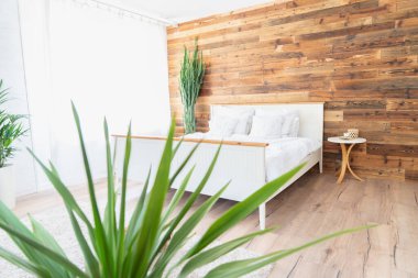 Boho tarzı rahat bir yatak odası. Büyük yatak, yeşil çiçekler ve ahşap duvar..