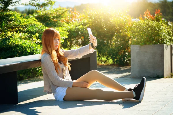 Ásia mulher sentado contra banco e selfie no telefone inteligente — Fotografia de Stock