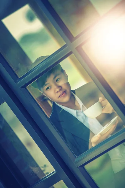 portrait of asia young businessman looking out window while standing in office and drinking coffee from cup, vintage effect