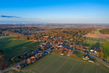 Açık mavi gökyüzünün altında yeşil tarlalar, ormanlar ve ormanlarla çevrili ağaçların arasında yuva yapan bir kasabanın insansız hava aracı görüntüsü.