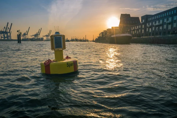 Floating buoy in a port with solar lantern to warn shipping of the channel boundaries with industrial cranes at a cargo depot silhouetted against a sunset and waterfront buildings