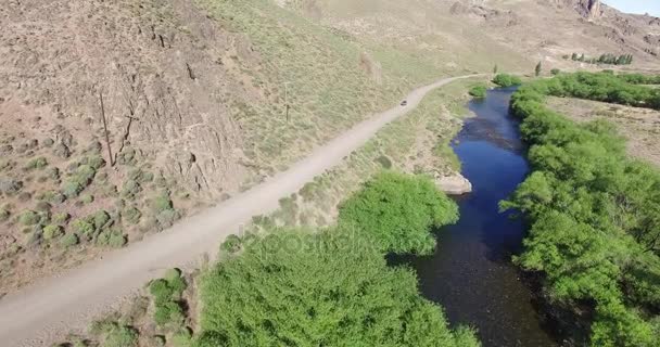 Drone aérien scène de route de gravier dans la steppe, van avancer contre caméra. Montagnes sur le fond. Paysage rural désertique avec rivière de pêche à la mouche et de grands arbres verts en Patagonie Argentine . — Video