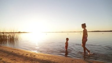 Kadın ve çocuk yavaş hareket ediyor, anne ve oğul, sahilde yürüyorlar altın saatinde suyun içinde. Kişilerin silueti. Cilt ışığı ve fişekler. Villa Soriano, Uruguay
