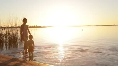 Kadın ve çocuk yavaş hareket ediyor, anne ve oğul, sahilde yürüyorlar altın saatinde suyun içinde. Kişilerin silueti. Cilt ışığı ve fişekler. Villa Soriano, Uruguay