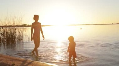 Kadın ve çocuk yavaş hareket ediyor, anne ve oğul, sahilde yürüyorlar altın saatinde suyun içinde. Kişilerin silueti. Cilt ışığı ve fişekler. Villa Soriano, Uruguay