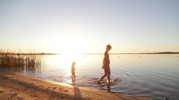 Zeitlupe von Frau und Kind, Mutter und Sohn, die in goldener Stunde am Strand entlang am Ufer im Wasser spazieren. Silhouette von Personen. Volumenlicht und Fackeln. villa soriano, uruguay — Stockvideo