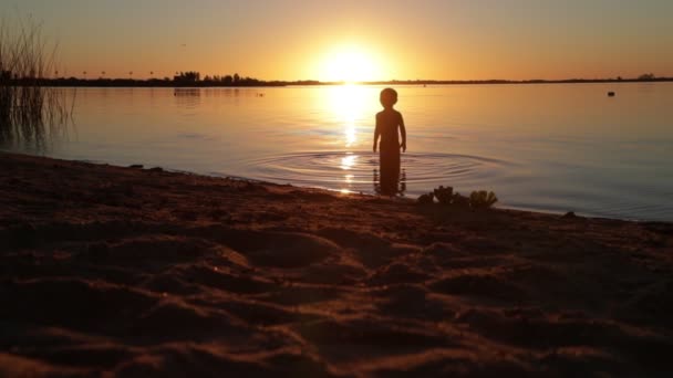 Zeitlupe eines glücklichen Kindes, das bei Sonnenuntergang aus dem Wasser in Richtung Sandstrand rennt. Sonne am Horizont versteckt, schwarze Silhouette der Person, goldene Stunde. villa soriano, uruguay — Stockvideo