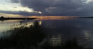 Çalılar ve nehir bitkilerinin üzerinde, turuncu günbatımını keşfeden ve yelkenlilerle kene yanaşan doğal bir kumsaldan uçan hava manzarası. Nehirdeki yansımalar. Villa Soriano, Uruguay