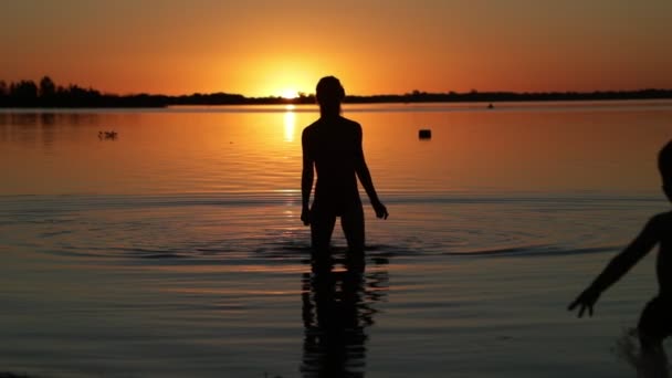 Αργή κίνηση σιλουέτας στο ηλιοβασίλεμα μητέρας και γιου να μπαίνουν στο νερό. Πορτοκαλί και μαύρη εικόνα. Ο ήλιος κρύβεται στον ορίζοντα. Villa Soriano, Ουρουγουάη — Αρχείο Βίντεο