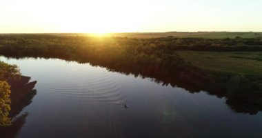Gün batımında sakin nehirde kanoyla kürek çeken birini takip eden hava sahnesi. Fişekler ve turuncu yansımalar nehirde. Arka planda güneş ve yeşil doğal manzara. San Salvador nehri, Dolores, Uruguay