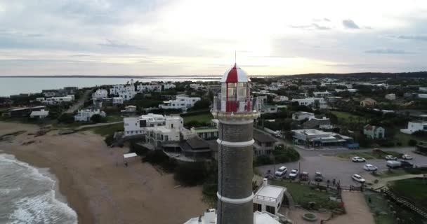 Cena aérea voando para trás mostrando detalhes do topo do farol descobrindo vista panorâmica da aldeia turística na península rochosa. José Ignacio, Maldonado, Uruguai — Vídeo de Stock