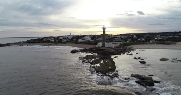 Letecký let z moře směrem k majáku a turistické vesnici na skalnatém poloostrově při západu slunce. Barevné barvy na pozadí. José Ignacio, Maldonado, Uruguay — Stock video