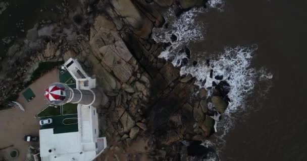 Luftaufnahme des Leuchtturms am Ufer. Wellen mit Schaumstoff, der im Hintergrund gegen Küstenfelsen spritzt. Steig- und Wendeschuss. jose ignacio, maldonado, uruguay — Stockvideo