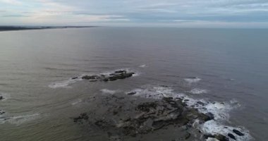Denizin üzerinden su yüzeyinden yükselen kayalara doğru uçan hava aracı görüntüsü. Dalgalar kayalık yapıların üzerine sıçrıyor. Jose Ignacio, Maldonado, Uruguay