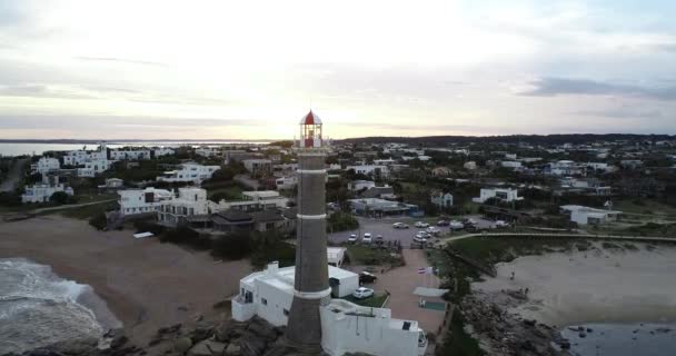 Primo piano aereo di camera lanterna, cupola e ponte galleria del faro. Volare verso e ruotare intorno alla costruzione. Città turistica e tramonto sullo sfondo. Jose Ignacio faro, Uruguay, Maldonado — Video Stock