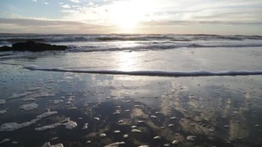 Gün batımında kumlu sahil şeridinde dalgaların yavaşça sıçraması. Köpük, kayalar ve su hareketi. Su yüzeyinde güneş yansıması. Jose Ignacio, Uruguay