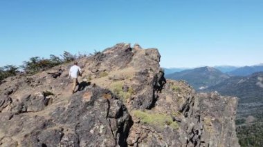 Uçan genç atletik adam kayalık zirveye ulaşıyor ve And Dağları Zinciri ve göllerin panoramik manzarasını keşfediyor. Parallax. Cerro Colorado yanardağına yolculuk, San Martin de los Andes, Neuquen