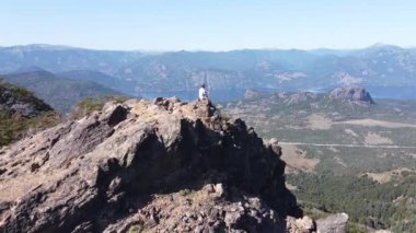 Kayalık Dağ Zirvesi 'nde, göllerin ve dağ zincirlerinin manzarasını izleyen genç bir adama doğru uçan hava aracı. Parallax. San Martin de los Andes, Neuquen, Arjantin