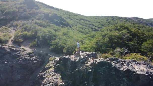 湖や山のチェーンの広い風景を見て山の岩の崖の上に若い陸上競技の男に関心のポイントを裏返しの航空機のドローンシーン。サン・マルタン・デ・ロス・アンデス,ノイケン,アルゼンチン — ストック動画