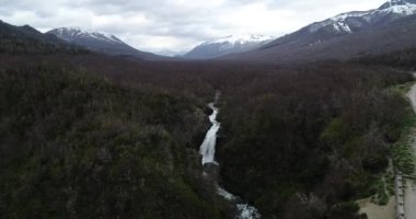 Hava aracı şelaleye doğru uçuyor. Tepeden tırnağa uçurumdan düşen suyun detaylarını gösteriyor. Yaprak döken orman ve arka planda karlı dağ zincirleri. Lanin Ulusal Parkı