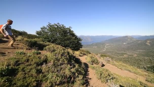 Cena de câmera lenta do jovem correndo descida ao longo da trilha rochosa e entrando na floresta. Lago Lacar e Corrente montanhosa dos Andes ao fundo. Patagônia, Argentina — Vídeo de Stock