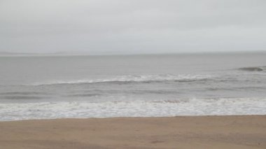 Kumlu sahilde dalgaların yavaş hareketi. Köpük ve su sıçratma detayı. Jose Ignacio kasabasında tatil yeri, Rocha, Uruguay. Sisli, bulutlu bir gün. Arkaplanda tepeler.