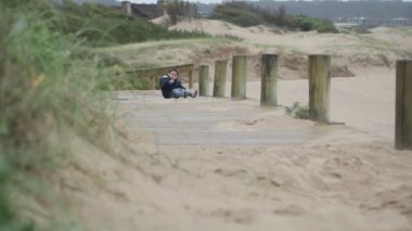 Kumsalda, tahta patikada oturan ve kumla oynayan bir çocuğun yavaş çekim sahnesi sahnenin sonunda ayağa kalkar. Arka planda kum tepeleri ve otlar. Jose Ignacio, Rocha, Uruguay