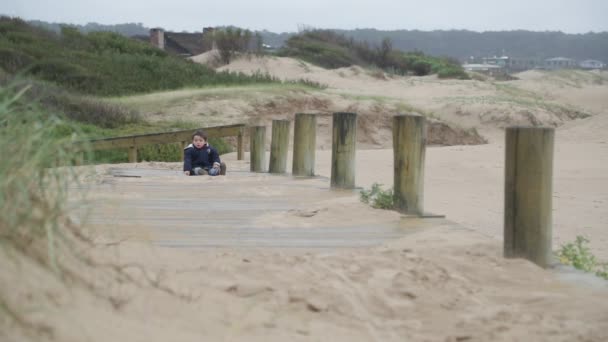 Медленная сцена движения ребенка на пляже, сидящего на деревянной дорожке. Песчаные дюны и травы на заднем плане. Хосе Игнасио, Роша, Уругвай — стоковое видео