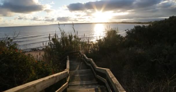 Går ner offentliga trä trappor mot stranden, solnedgång och havsvågor i bakgrunden. Solis, Uruguay — Stockvideo