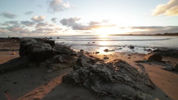 Slow motion of beach at golden hour. Foreground of rocks at shoreline while waves brake at the sand. Golden reflection over sea surface. Sunset at the horizon. Piriapolis, Uruguay — Stock Video