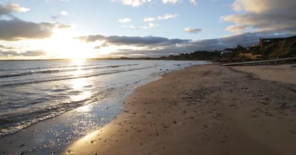 ビーチ黄金の時間で、海岸線に沿って歩く一方、波の砂でブレーキ。海面上の黄金の反射。地平線の夕日。ウルグアイのピリオポリス — ストック動画