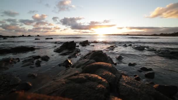 Langzame beweging van het strand op het gouden uur. Voorgrond van rotsen aan de kust, terwijl golven breken op het zand. Gouden reflectie over het zeeoppervlak. Zonsondergang aan de horizon. Piriapolis, Uruguay — Stockvideo