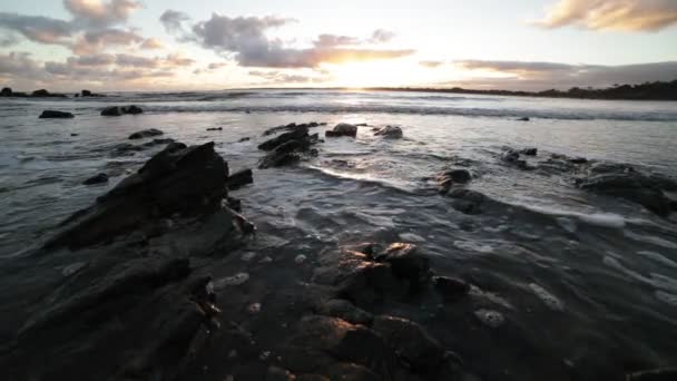 Movimiento lento de la playa a la hora dorada. Primer plano de las rocas en la costa, mientras que las olas frenan en la arena. Reflejo dorado sobre la superficie del mar. Puesta de sol en el horizonte. Piriapolis, Uruguay — Vídeos de Stock